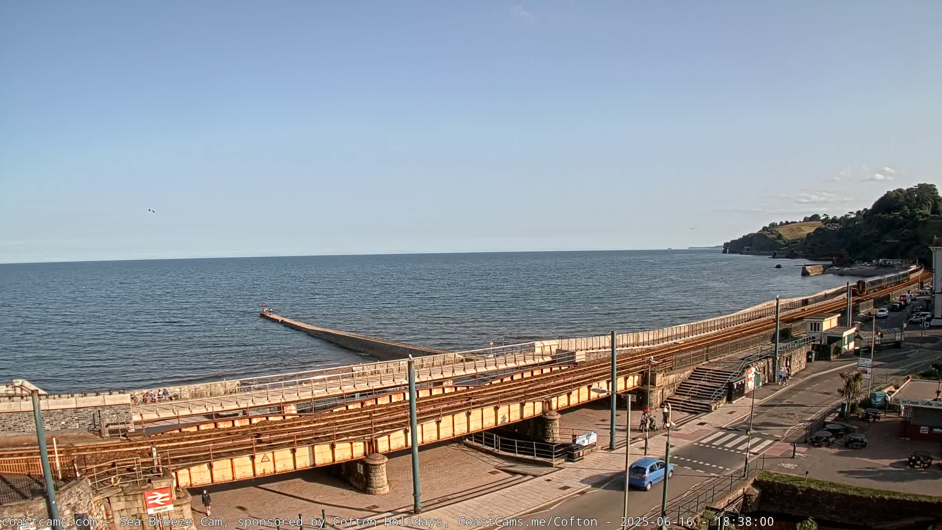 A train track runs alongside a calm ocean, passing over a stone structure that extends from the shore into the water under a clear, sunny sky.