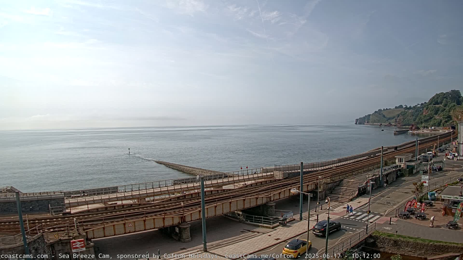 A coastal scene on a partly sunny day shows a railway line crossing a bridge over a waterfront area, with calm sea waters extending to a hazy horizon and green hills in the background.