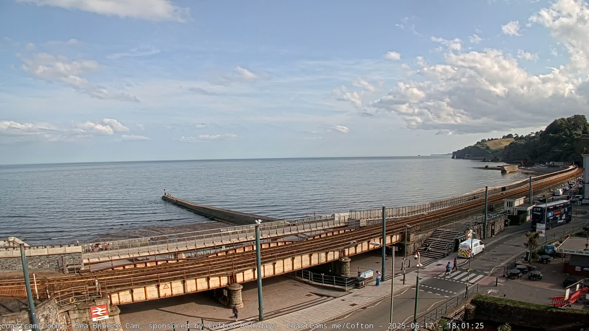 A partly cloudy day shows a coastal town scene with a railway line crossing a seaside promenade that leads to a breakwater extending into a calm sea.