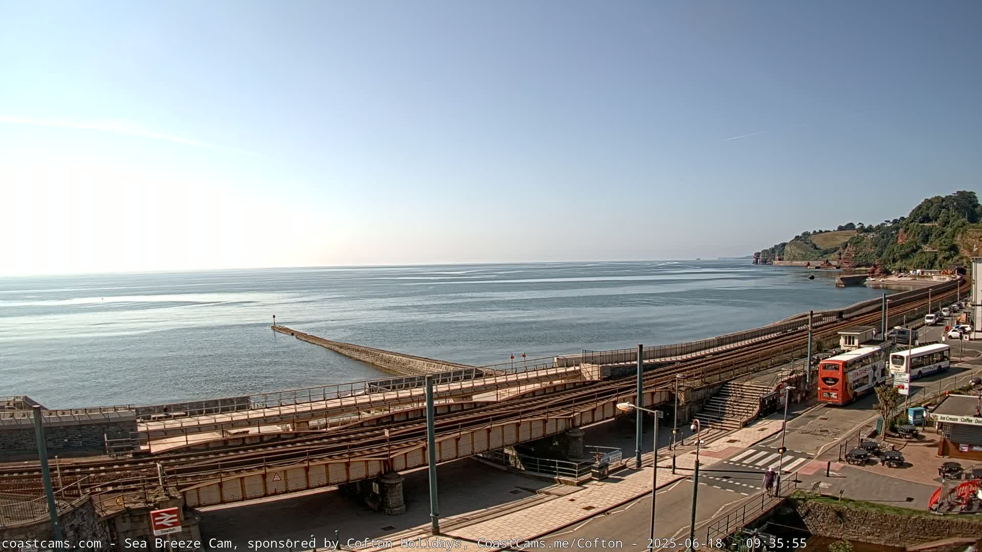 A calm, sunny day reveals a coastal scene with train tracks leading to a harbor, a breakwater extending into the sea, and buses parked near a building.