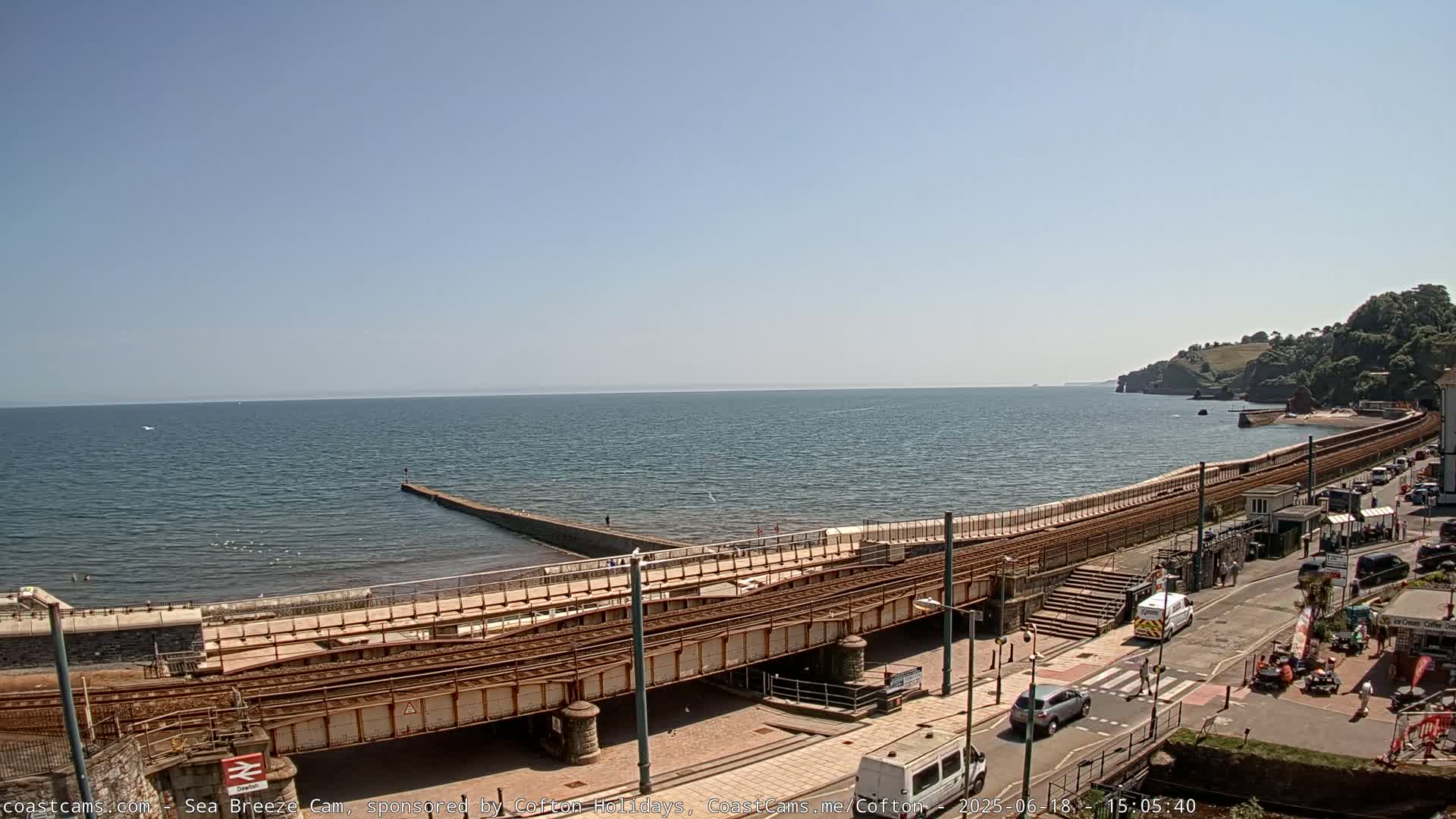 A seaside scene on a sunny day shows a railway line running along a sea wall, with a calm ocean in the background and a few cars parked on a street nearby.