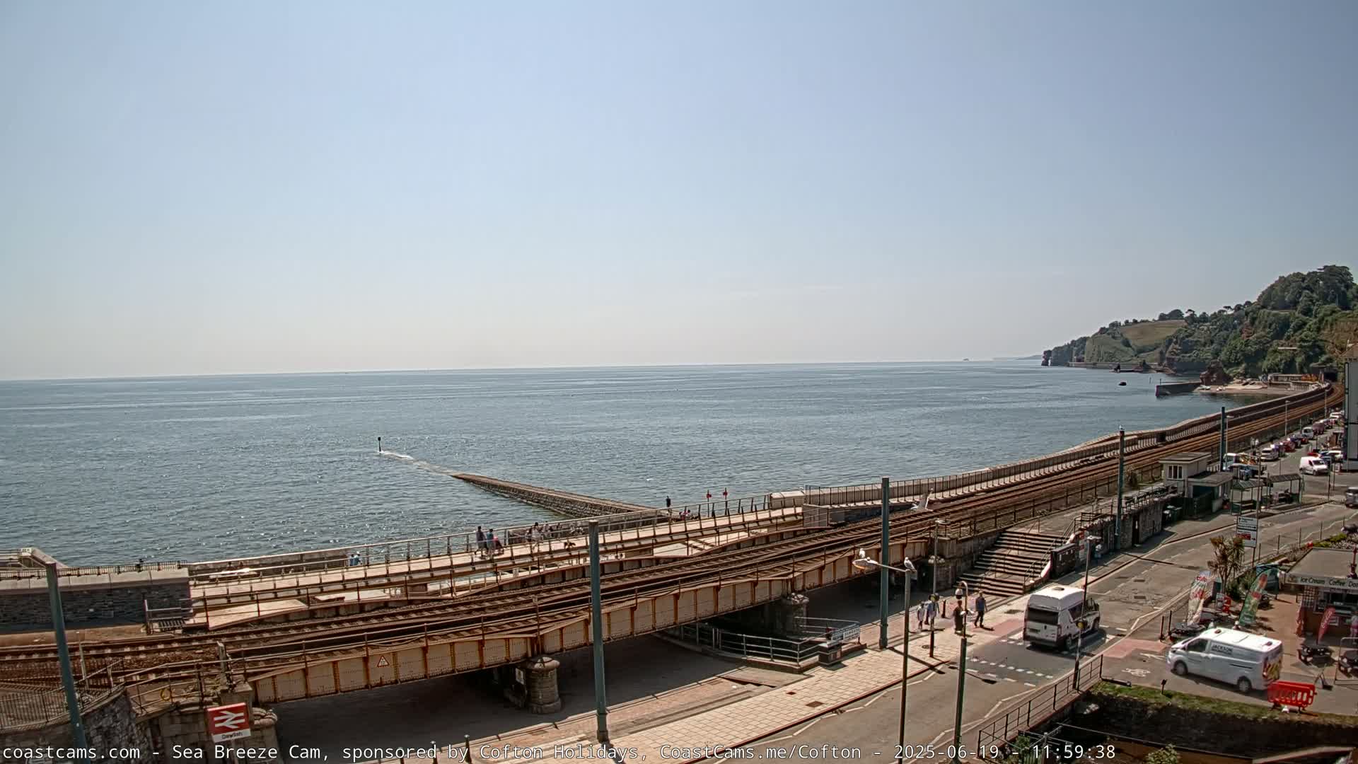 A coastal scene on a sunny day shows train tracks running along a sea wall next to a road with vehicles and pedestrians, overlooking a calm ocean.