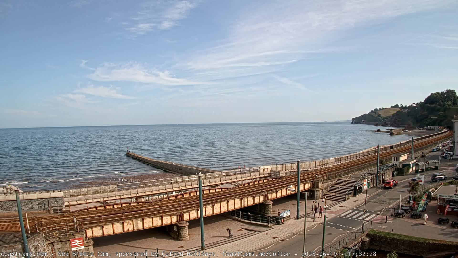 A seaside town scene on a partly sunny day shows a railway line crossing a beach, leading to a calm ocean with a distant coastline.