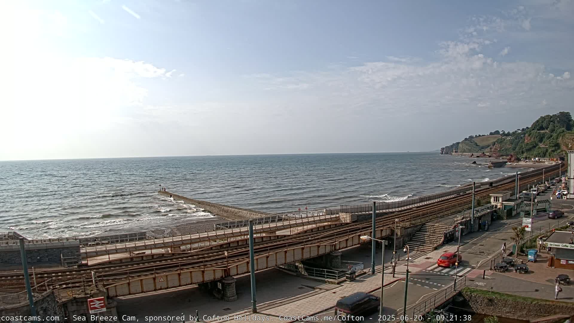 A mostly sunny day shows a coastal scene with a railway line running alongside a beach where small waves are breaking, meeting a low sea wall.