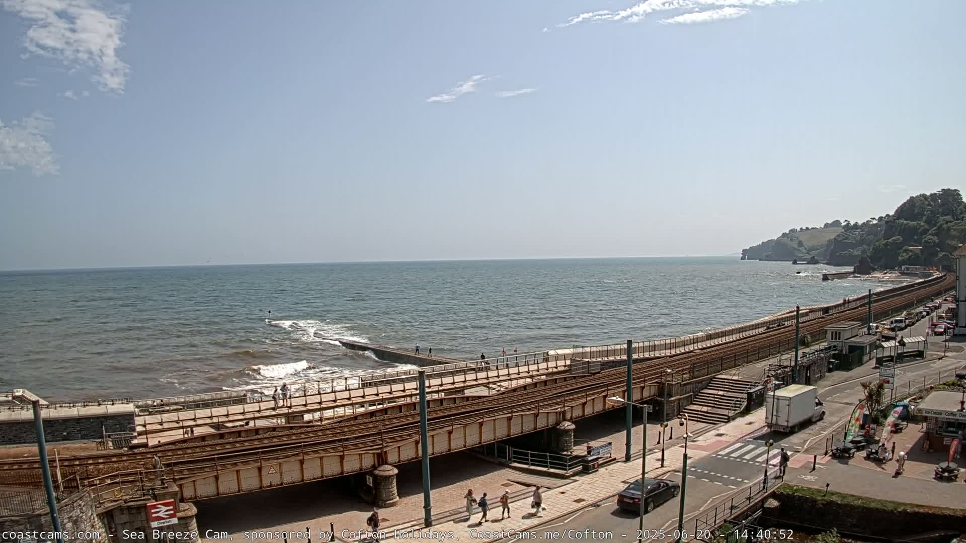 A mostly sunny day shows a seaside town with a railway track running alongside a shoreline, gentle waves breaking on the shore, and some people walking along the path.