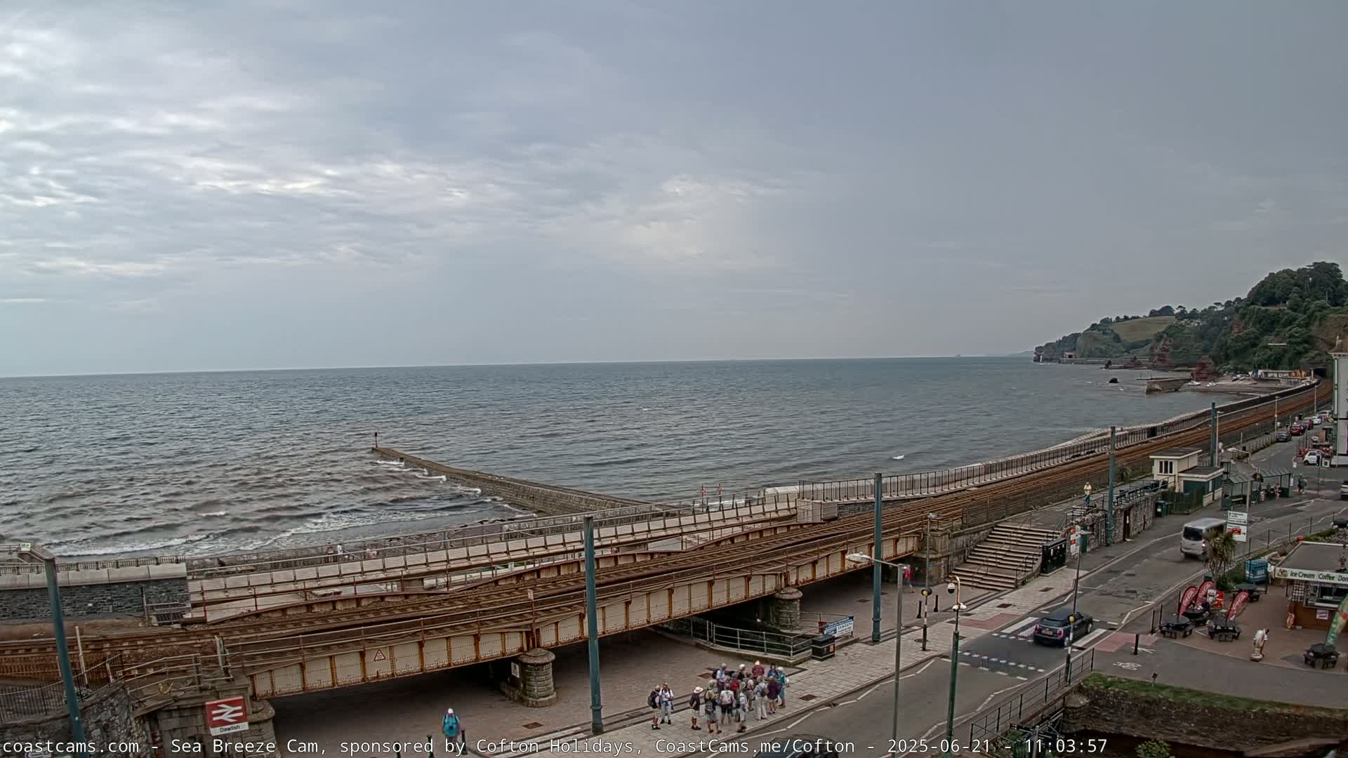 A coastal scene under a cloudy sky shows a train track crossing over a road next to a beach with moderate waves and a rocky coastline in the distance.