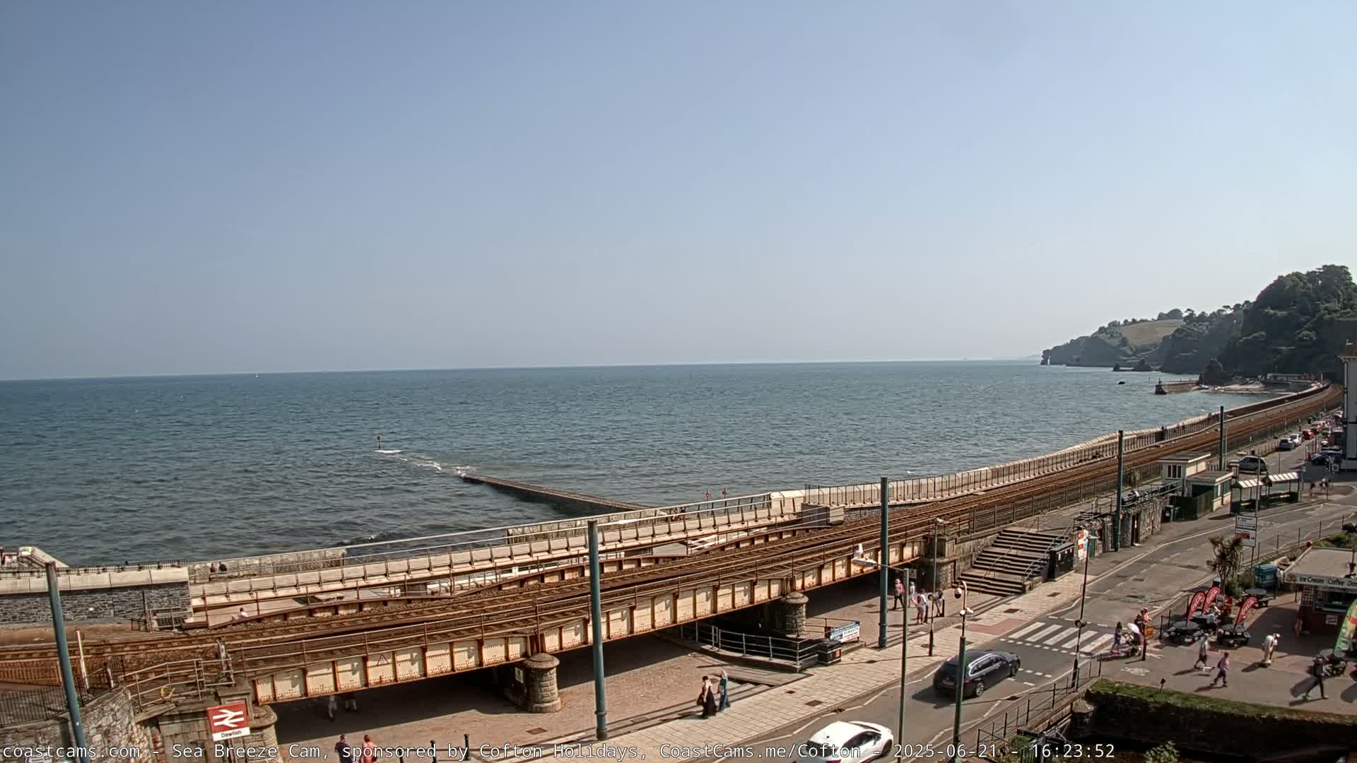 A seaside town on a sunny day shows a railway line running alongside a sea wall and a beach with people walking nearby.