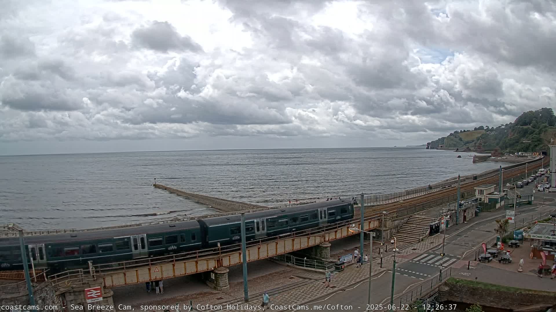 A dark-green train crosses a bridge over a coastal road, under a cloudy sky, beside a wavy sea with a breakwater.