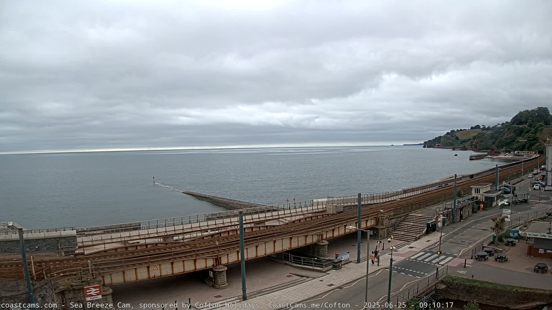A railway bridge crosses a coastal road next to a calm sea under an overcast sky.