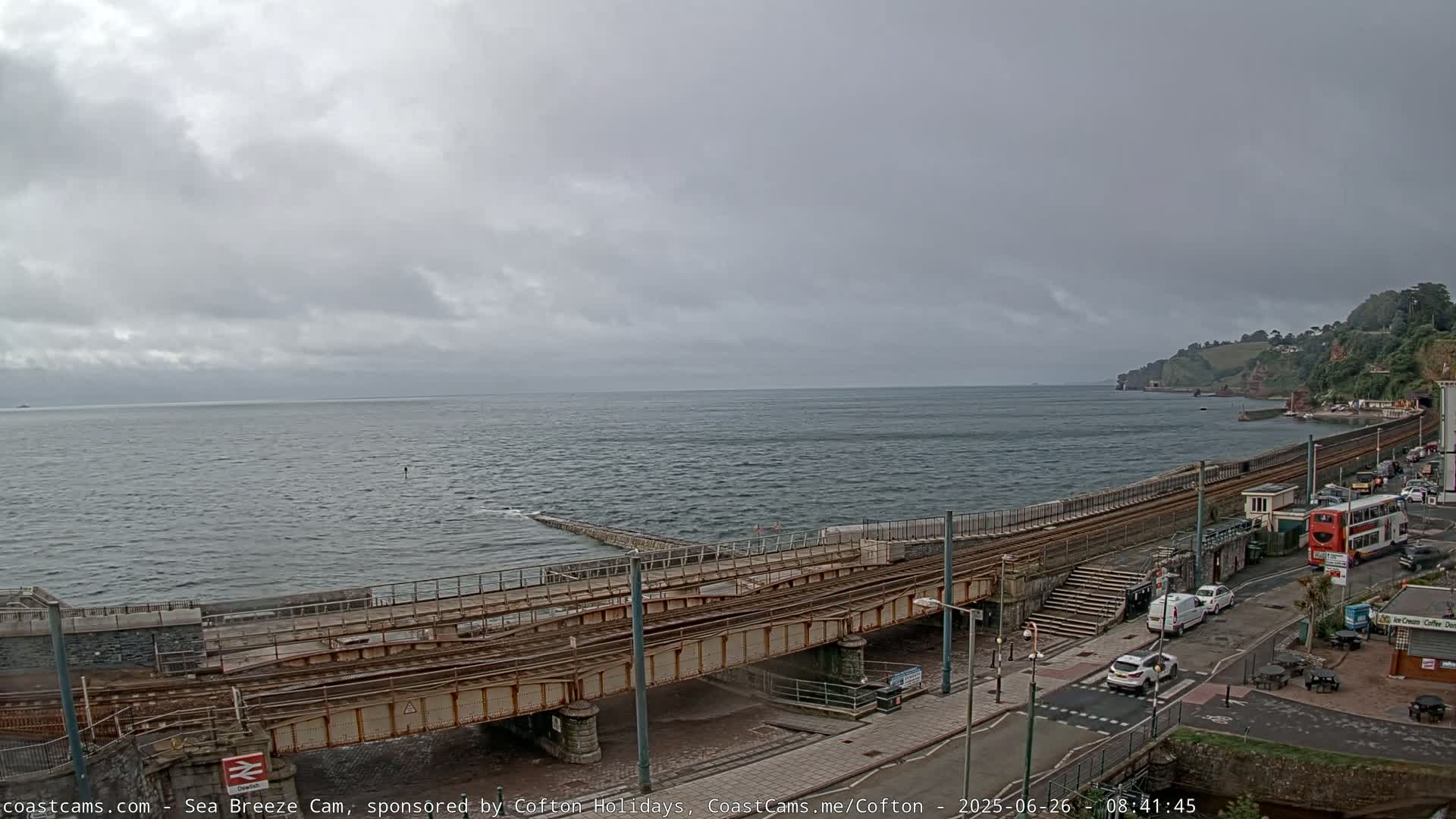 A railway track crosses a coastal road beside a choppy sea under an overcast sky.