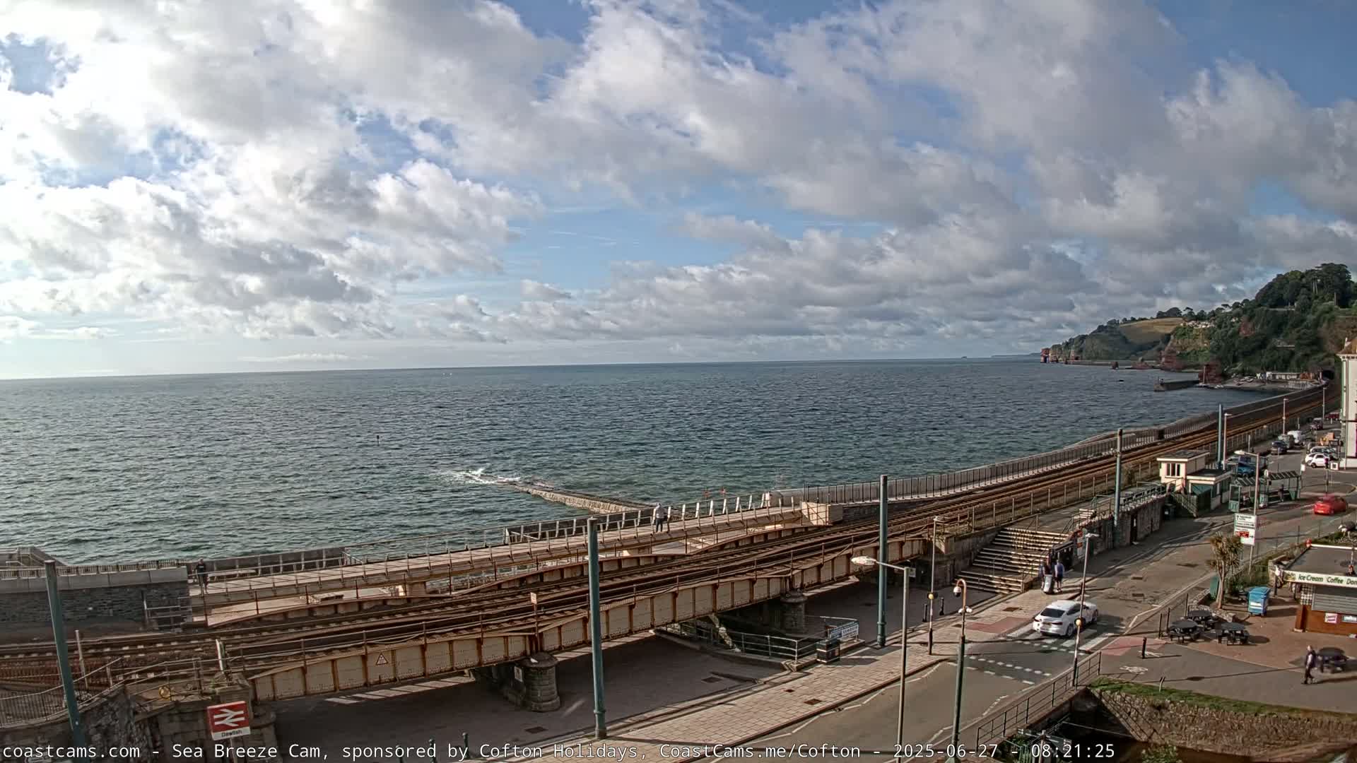 A coastal scene under a partly cloudy sky shows a railway line running alongside a seawall, with a road and buildings visible beyond.