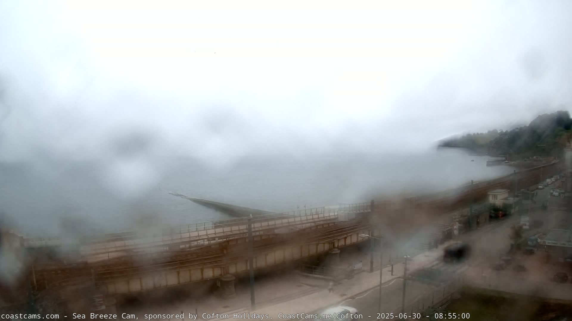 A rain-streaked view shows a coastal scene with a sea wall, a roadway, and some buildings, all obscured by fog and heavy rain.