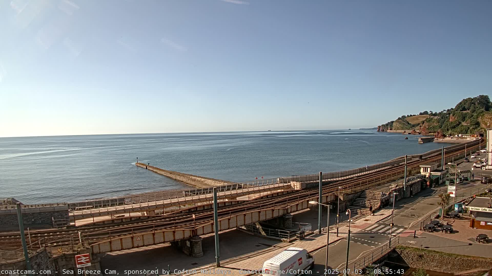 A calm sea meets a shoreline with train tracks and a seaside town under a clear, sunny sky.