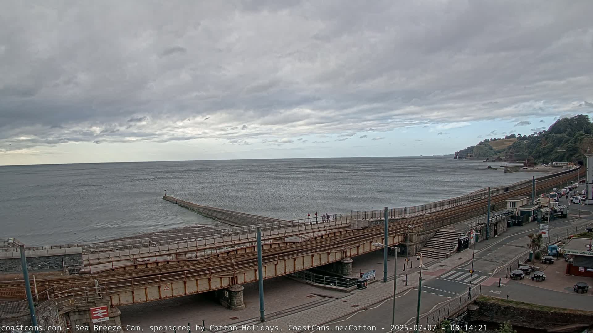 A coastal scene under a cloudy sky shows a railway line running along a beach, with a sea wall and a small pier extending into the water.