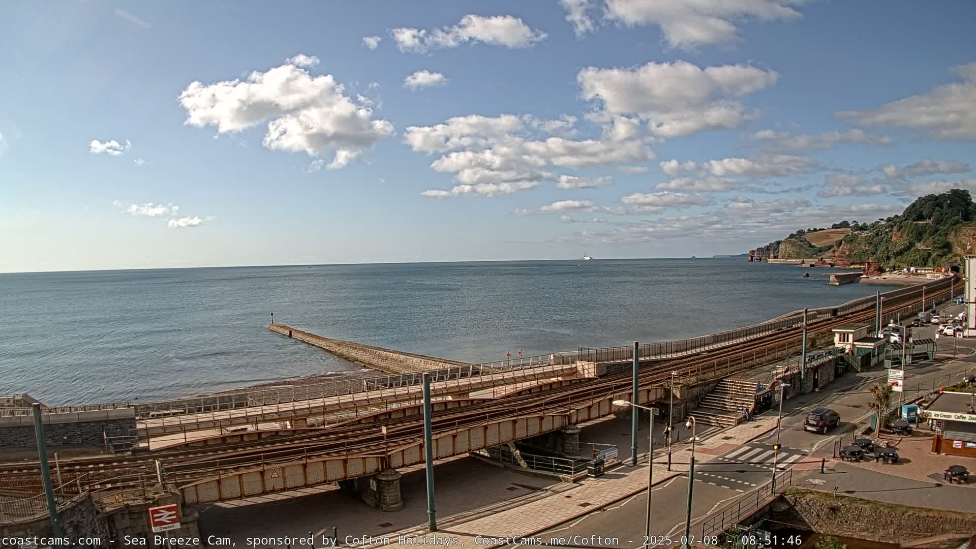 A partly cloudy day shows a coastal scene with a train track crossing a beach, a small pier extending into the sea, and a shoreline with buildings.