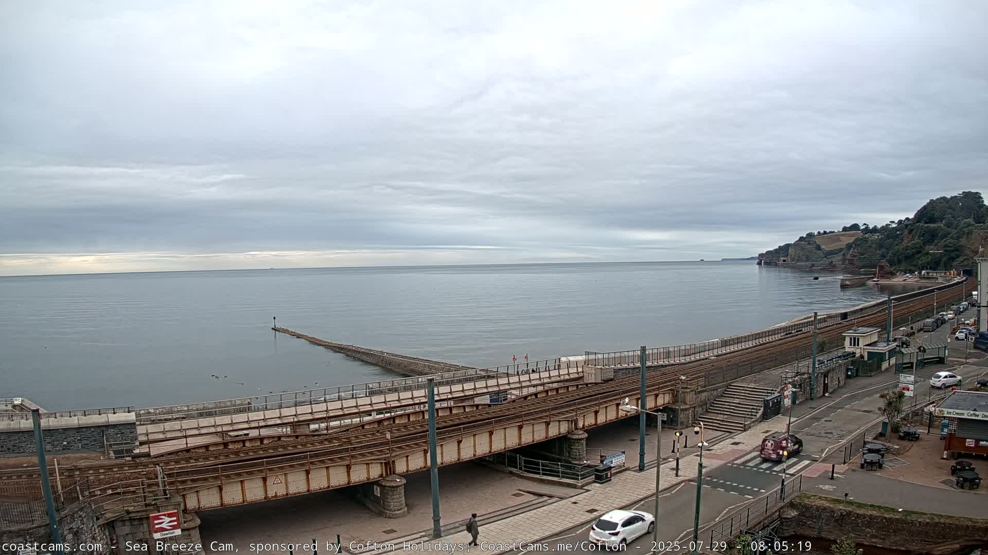 A railway bridge crosses a coastal road next to a calm sea under an overcast sky.