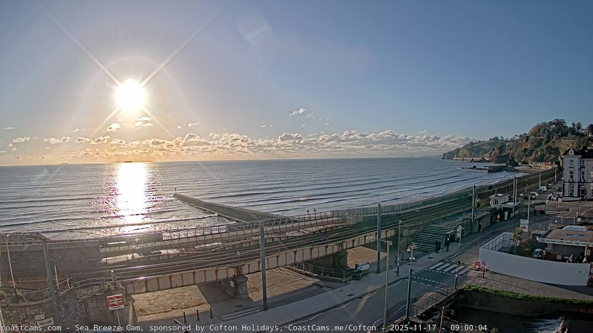 Under a bright, sunny sky with scattered clouds, a coastal landscape features a sun-drenched sea reflecting the low sun, a long pier, a railway line running along the beach, and a road with buildings at the base of rocky, green cliffs.