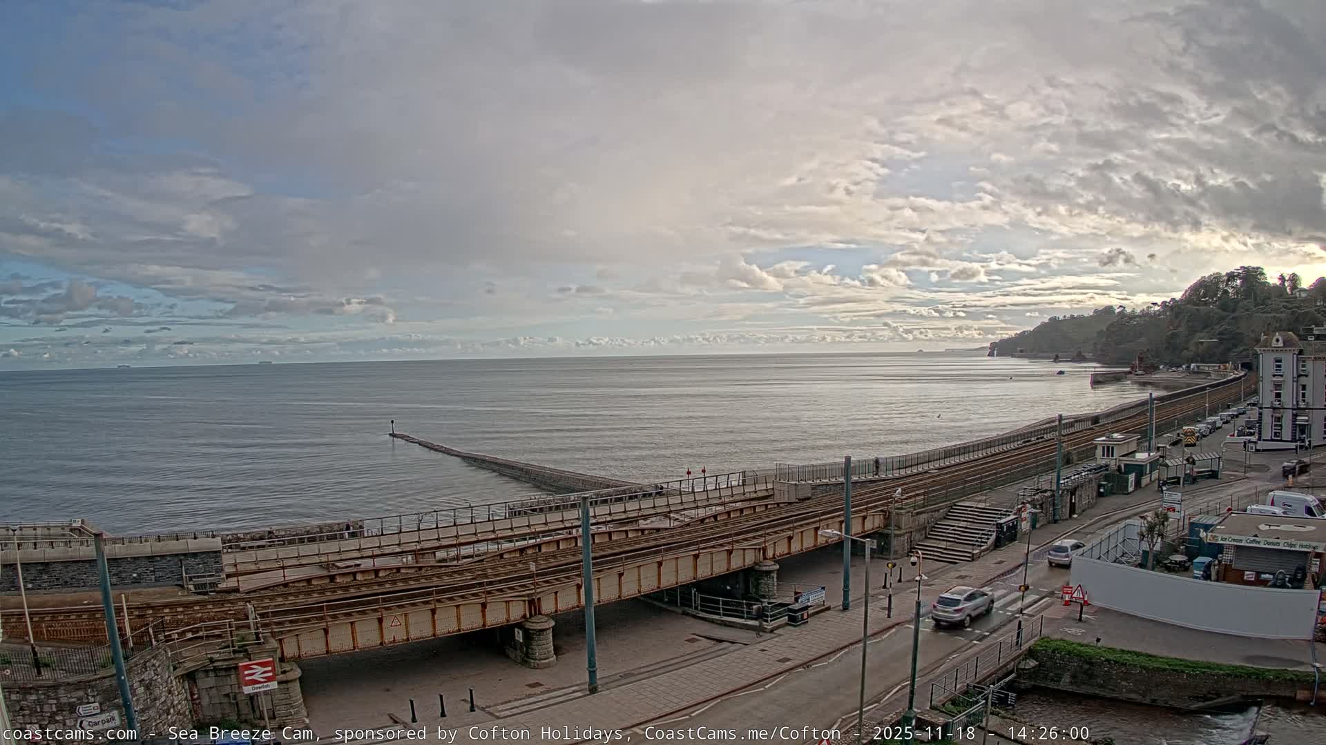 Under a bright, sunny sky with scattered clouds, a coastal landscape features a sun-drenched sea reflecting the low sun, a long pier, a railway line running along the beach, and a road with buildings at the base of rocky, green cliffs.