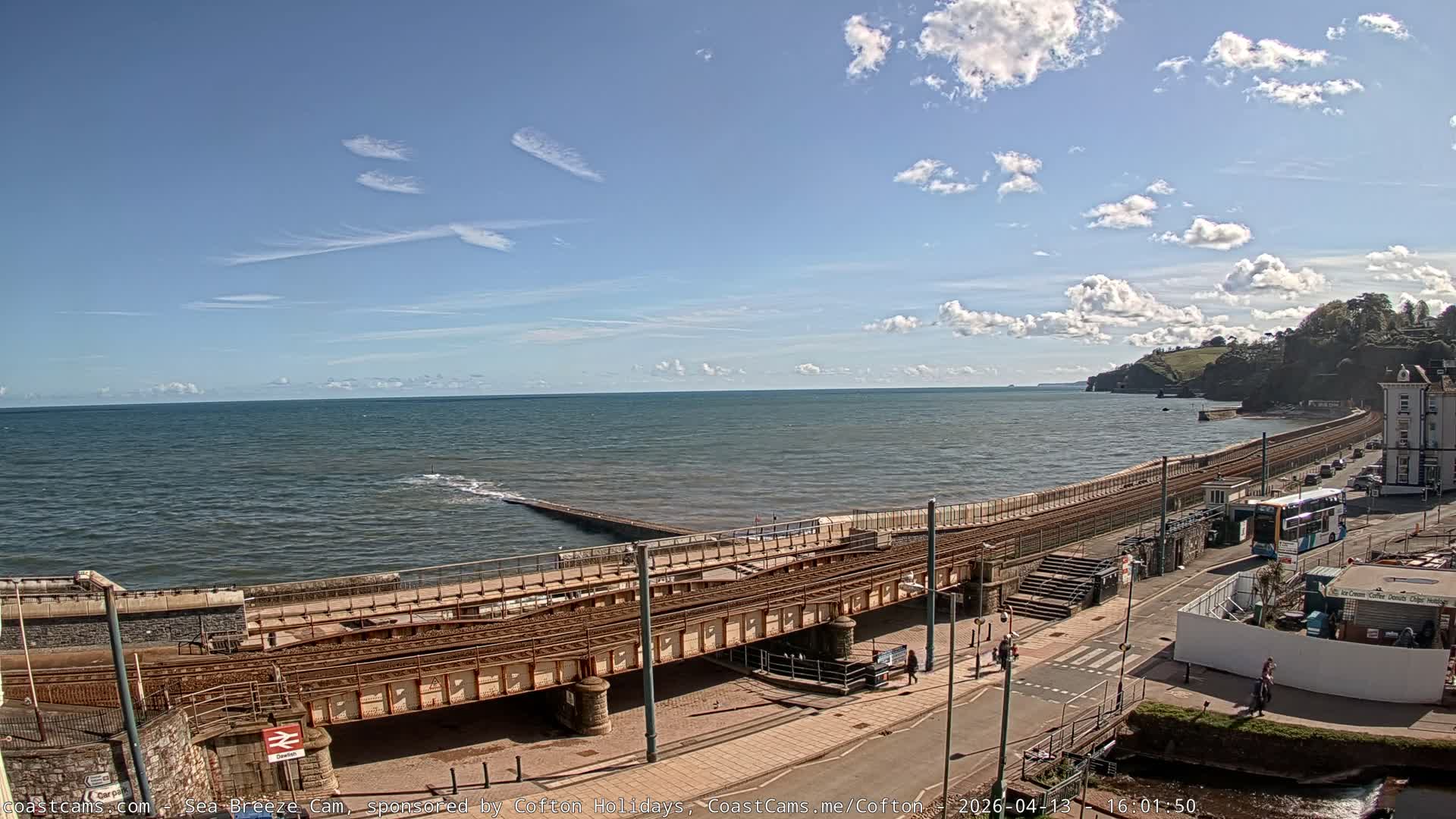 Under a bright, sunny sky with scattered clouds, a coastal landscape features a sun-drenched sea reflecting the low sun, a long pier, a railway line running along the beach, and a road with buildings at the base of rocky, green cliffs.