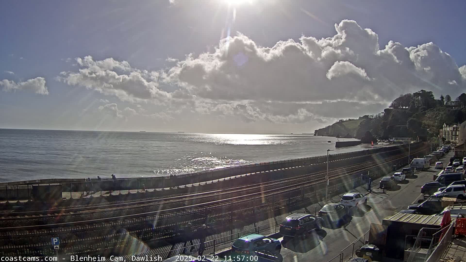 A partly sunny day at the coast shows a calm sea, a railway line running along a seawall, and cars parked along a road next to green cliffs.