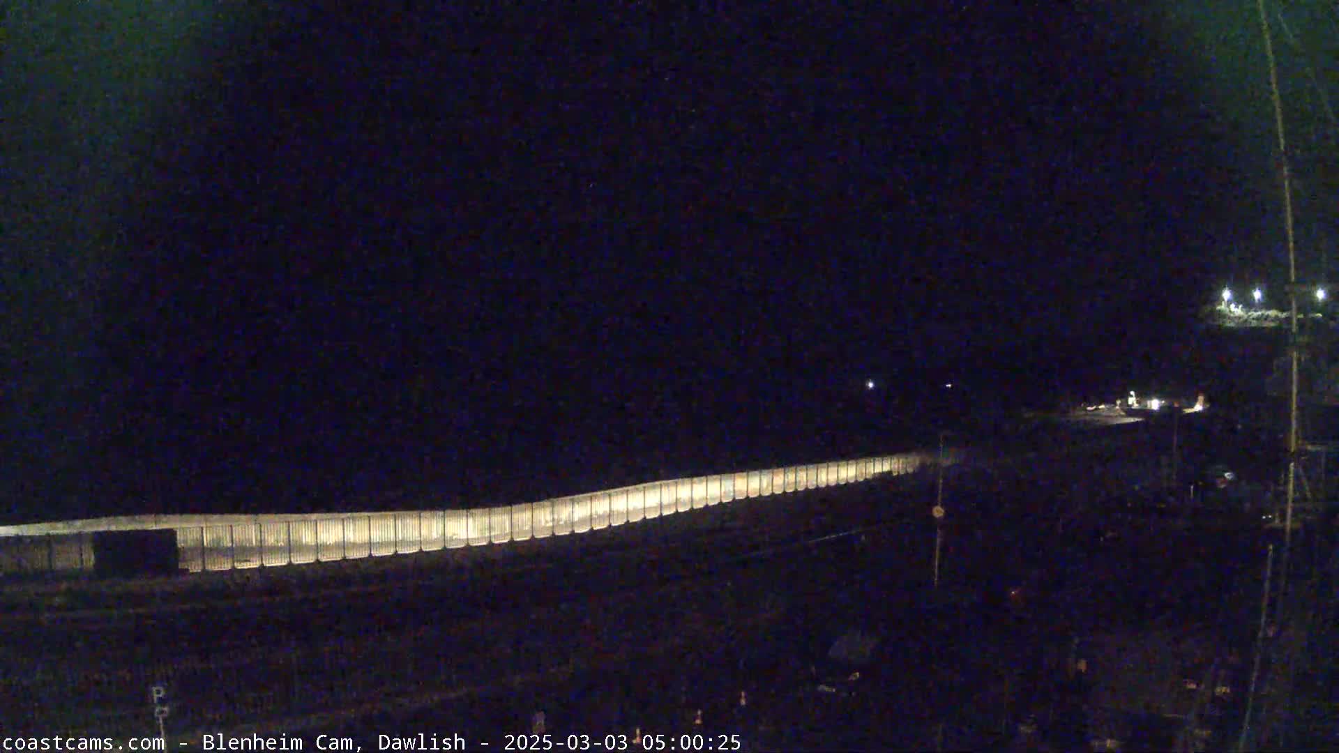 A long, illuminated fence stretches across a dark, nighttime scene, with some vehicles visible in the distance.