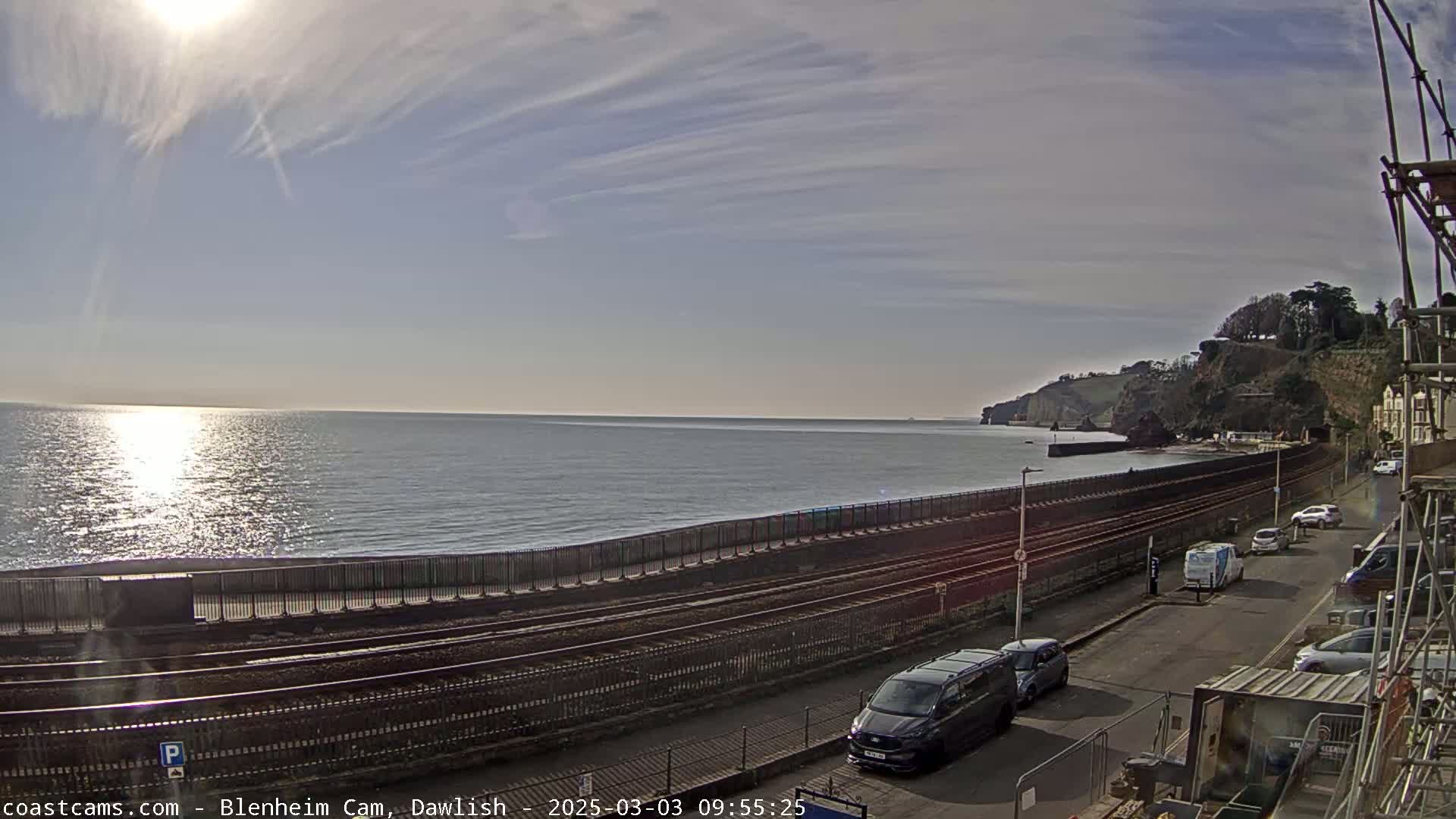 A sunny day with a partly cloudy sky overlooks a calm sea, railroad tracks, a road with parked cars, and a cliffside with vegetation.
