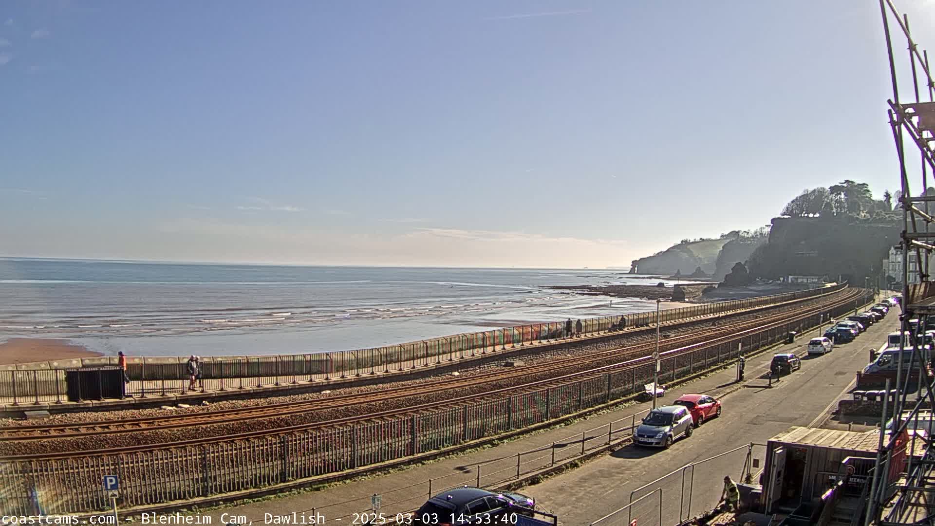 A railway track runs along a beach with a calm sea under a clear blue sky, and cars are parked along a road next to the tracks.