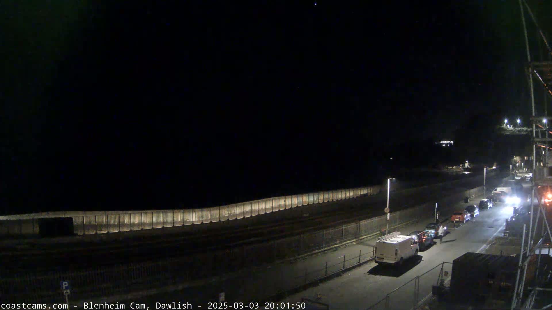 A nighttime view shows a seawall, a road with several parked cars, and a clear, dark sky.