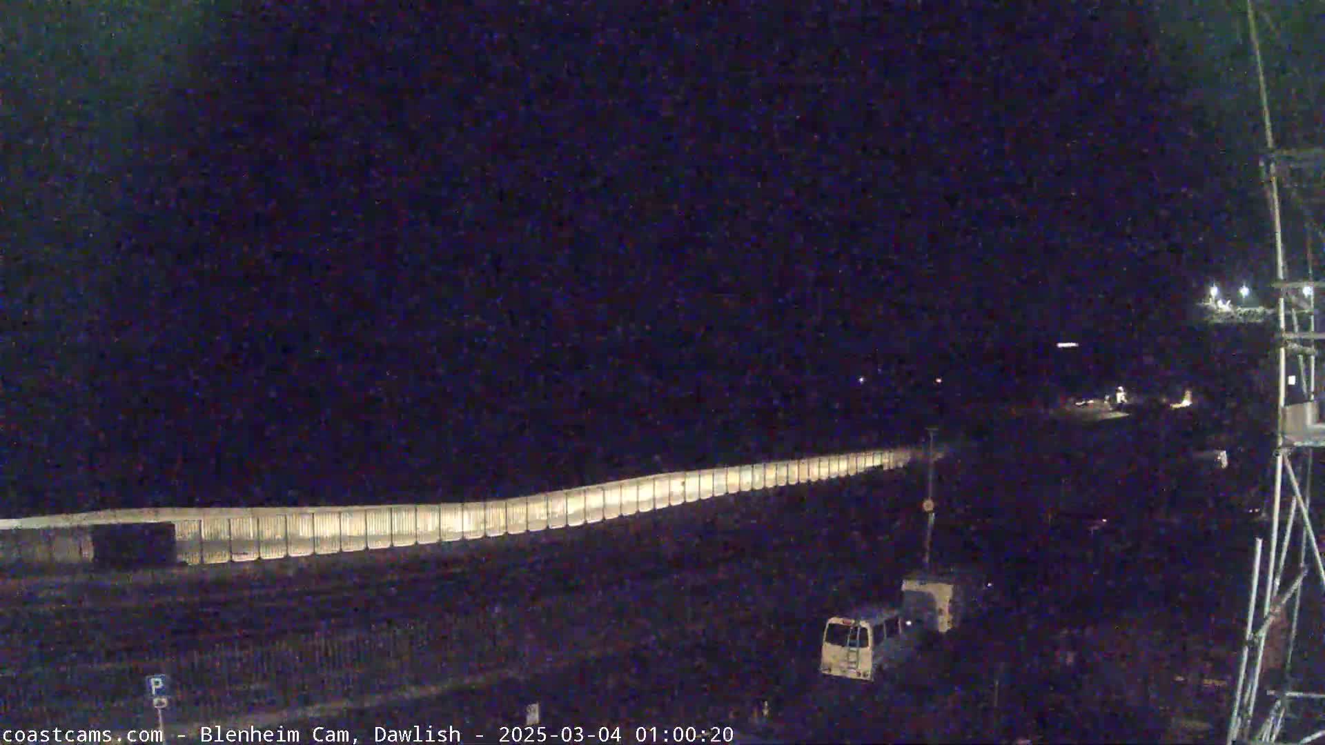 A long, illuminated barrier stretches across a dark, nighttime scene, with a few vehicles visible near a tall metal structure.