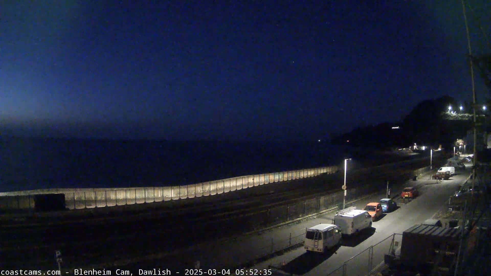 A dark, nighttime coastal scene shows a long wall separating a road with parked vehicles from a calm sea under a clear, dark-blue sky.