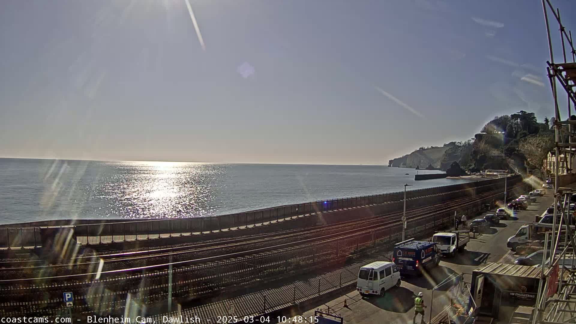 A sunny day shows a calm ocean, a railway track next to a seaside road with parked cars, and cliffs in the background.