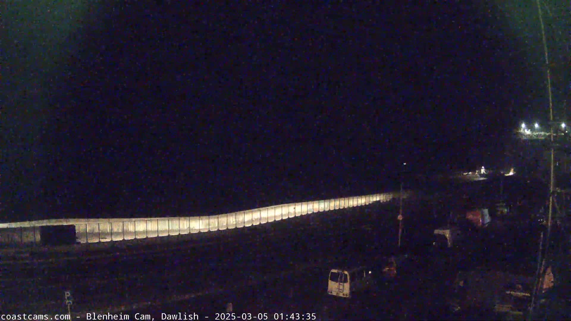 A long, illuminated fence stretches across a dark, nighttime scene, with a few vehicles visible in the lower right corner.
