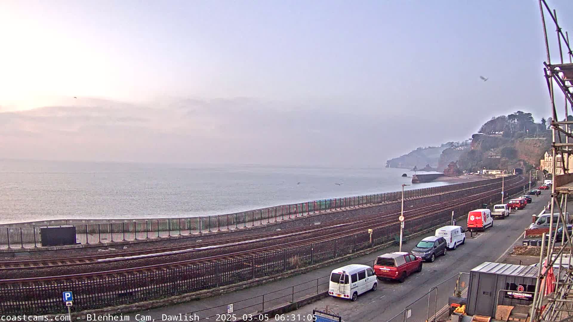 A calm sea stretches to a hazy horizon under a pale sky, with railway tracks and parked vehicles lining a coastal road beside red cliffs.