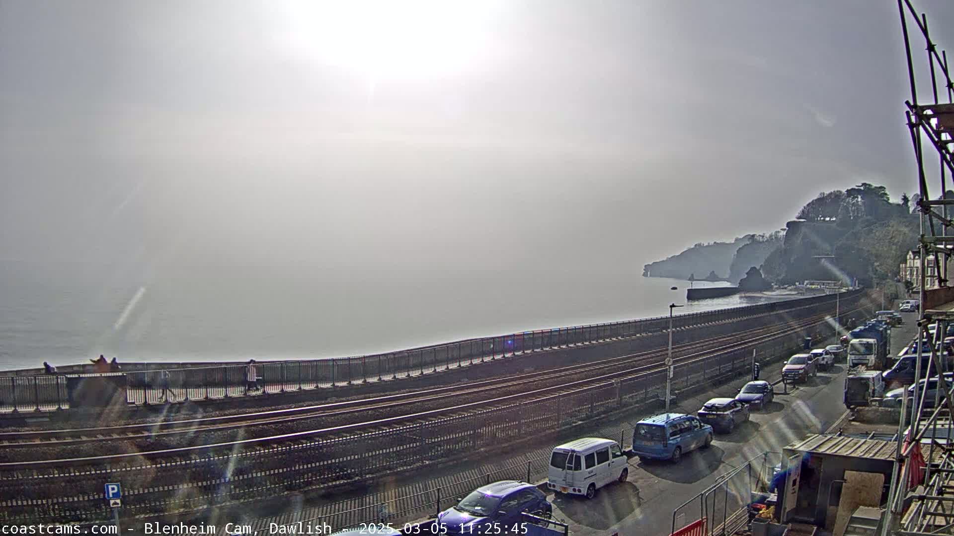 A hazy, sunny day reveals a coastal scene with railroad tracks running alongside a seawall, a parked line of cars, and cliffs in the background.