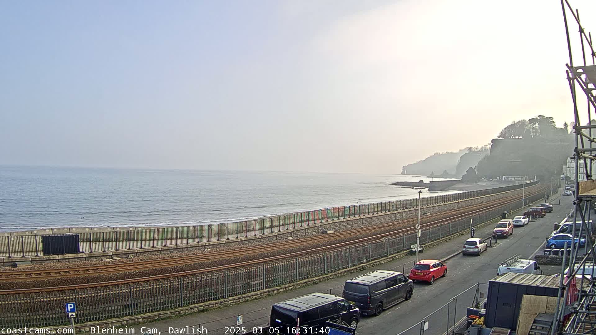 A hazy, sunny day overlooks a calm ocean with a beach, seawall, train tracks, and parked cars along a road next to cliffs.