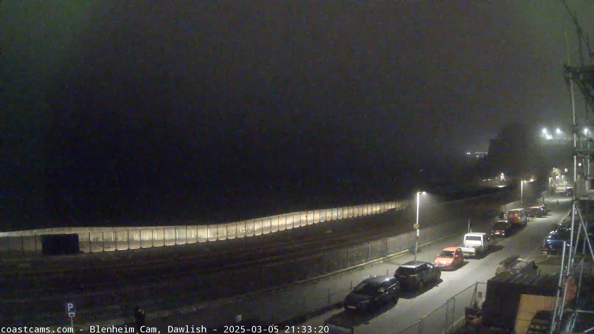 A foggy night scene shows several cars parked along a road next to a tall, illuminated wall.