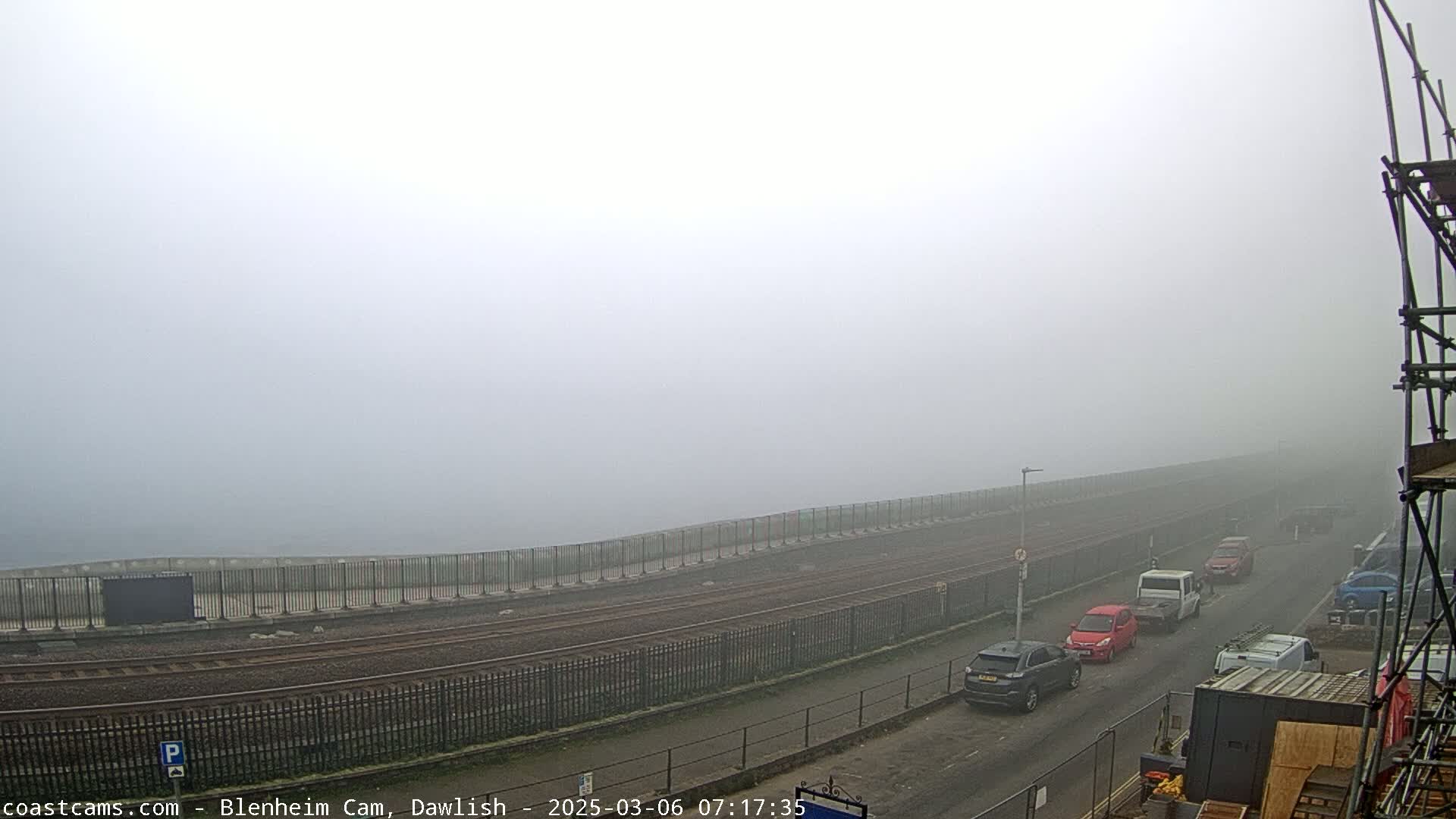A foggy scene shows a roadway next to train tracks, with several cars parked along the road and scaffolding in the bottom right corner.