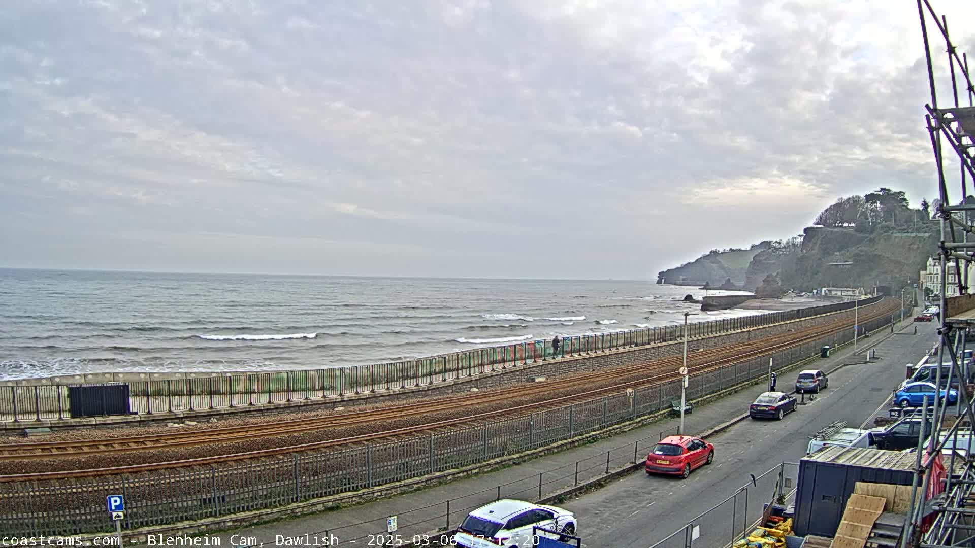 A mostly cloudy day shows a sea wall and railway line alongside a beach with small waves, overlooking a rocky cliffside.