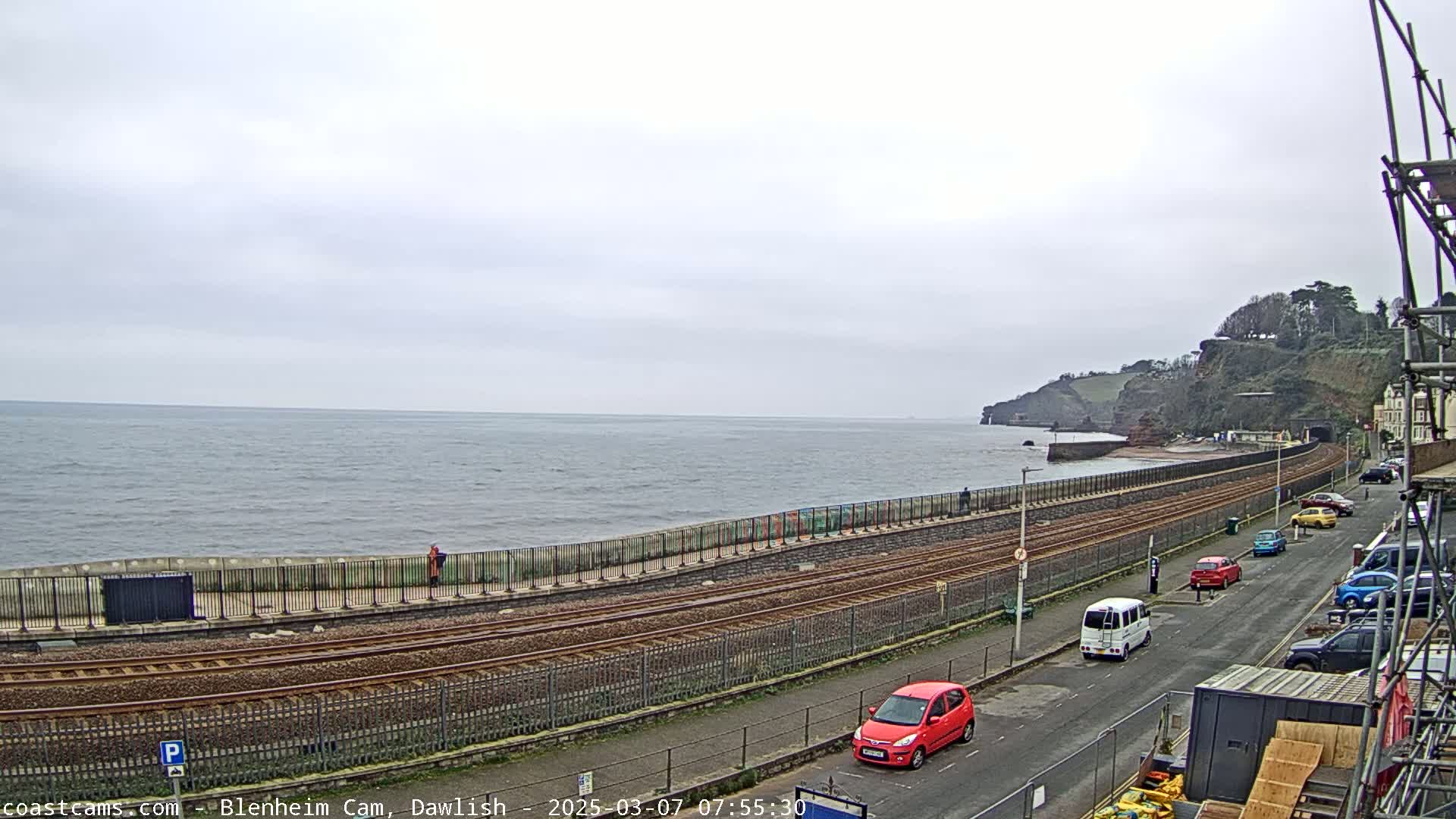 A coastal railway line runs alongside a seawall, with parked cars and a few pedestrians visible on an overcast day.