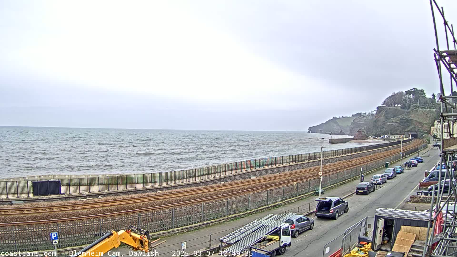 A choppy sea stretches to a cliffside under an overcast sky, with railway tracks running alongside a road lined with parked cars and construction equipment.