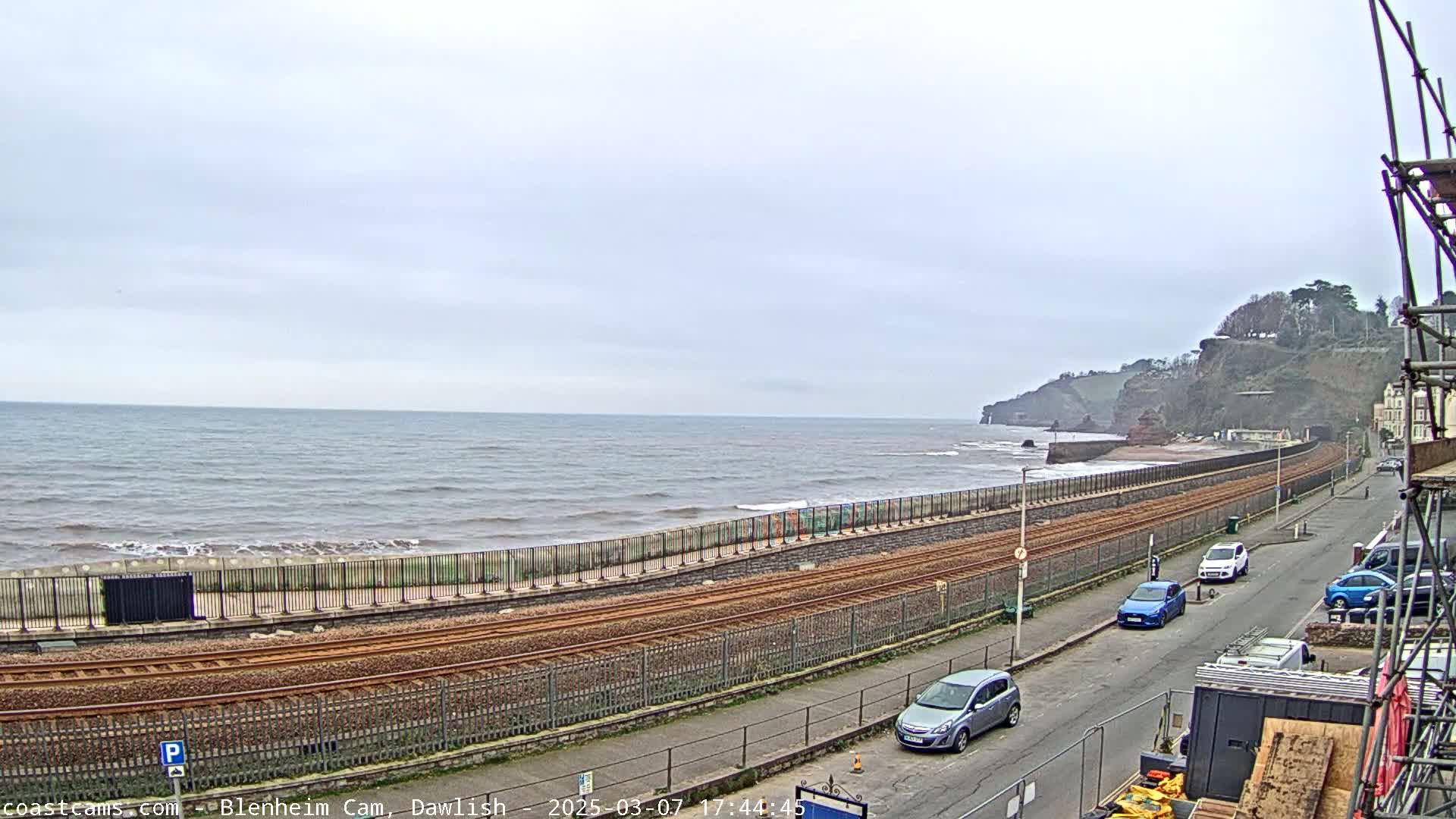 A coastal scene on an overcast day shows train tracks running alongside a sea wall, with cars parked on a road next to it, all overlooking a calm sea and cliffs in the distance.