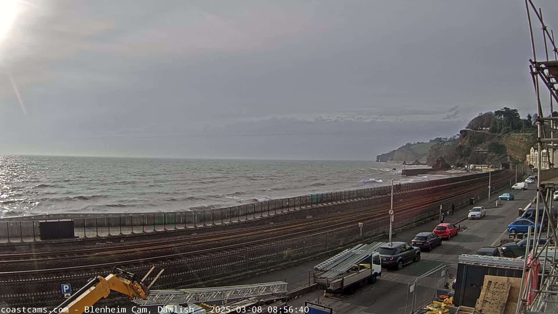 A choppy sea with waves breaking against a seawall next to a railway line and a street with parked cars, under a mostly cloudy sky.