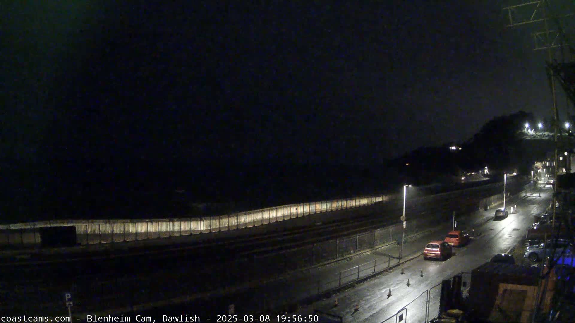 A nighttime view of a wet road next to a seawall, with a few cars visible under dark, overcast skies.