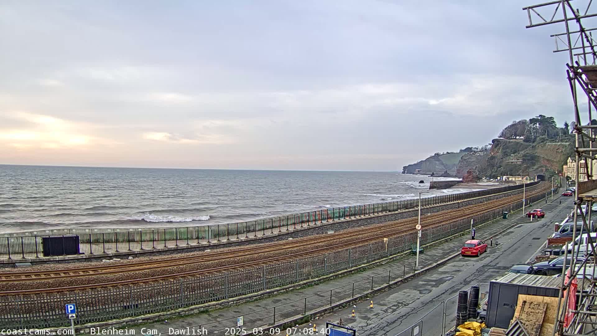 A coastal railway line runs alongside a sea wall, with a somewhat overcast sky and small waves breaking on the shore.