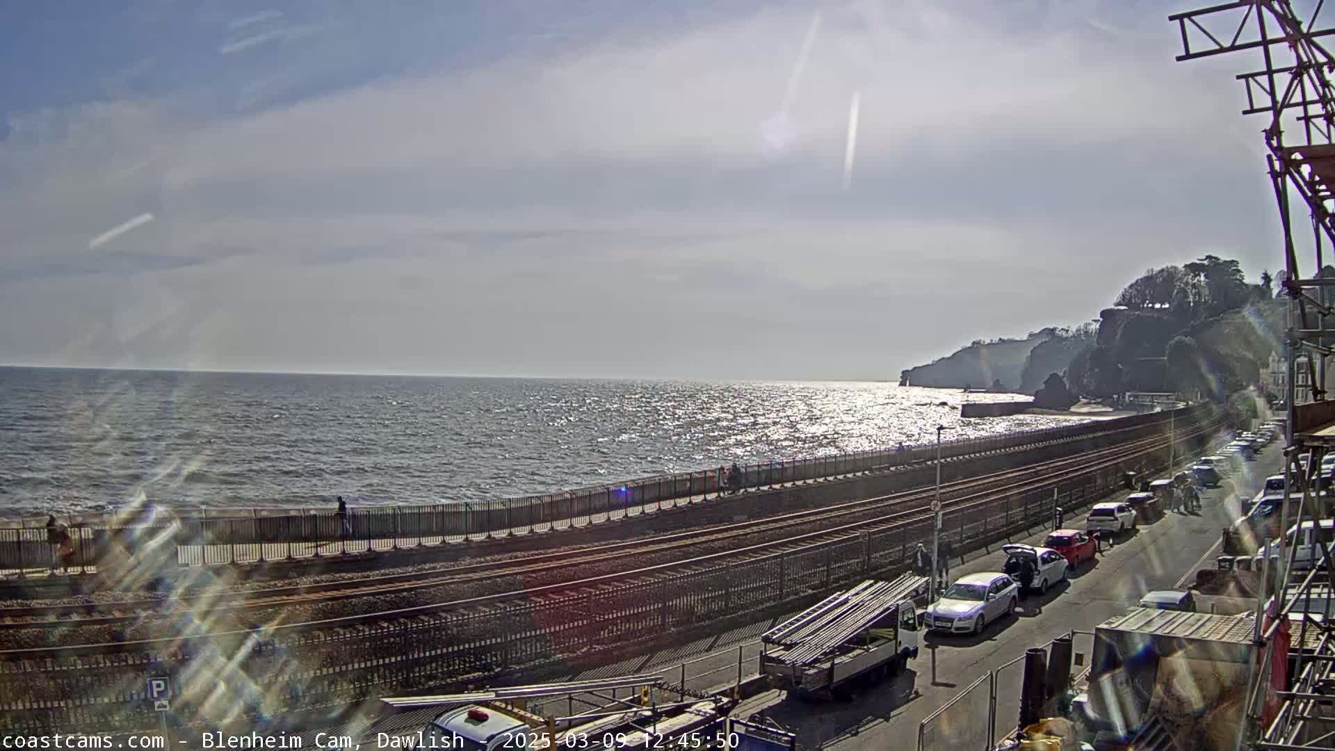 A mostly sunny day shows a coastline with a train track running along a paved road next to the ocean, where several cars are parked and a few people walk.