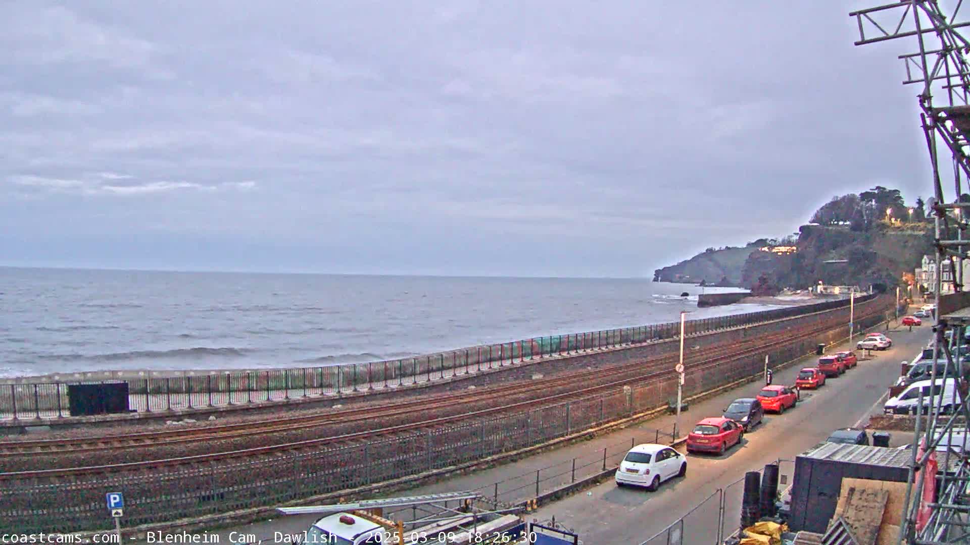 A coastal scene under an overcast sky shows a train track running parallel to a seawall with parked cars along a road beyond it, and a rocky cliff in the distance.