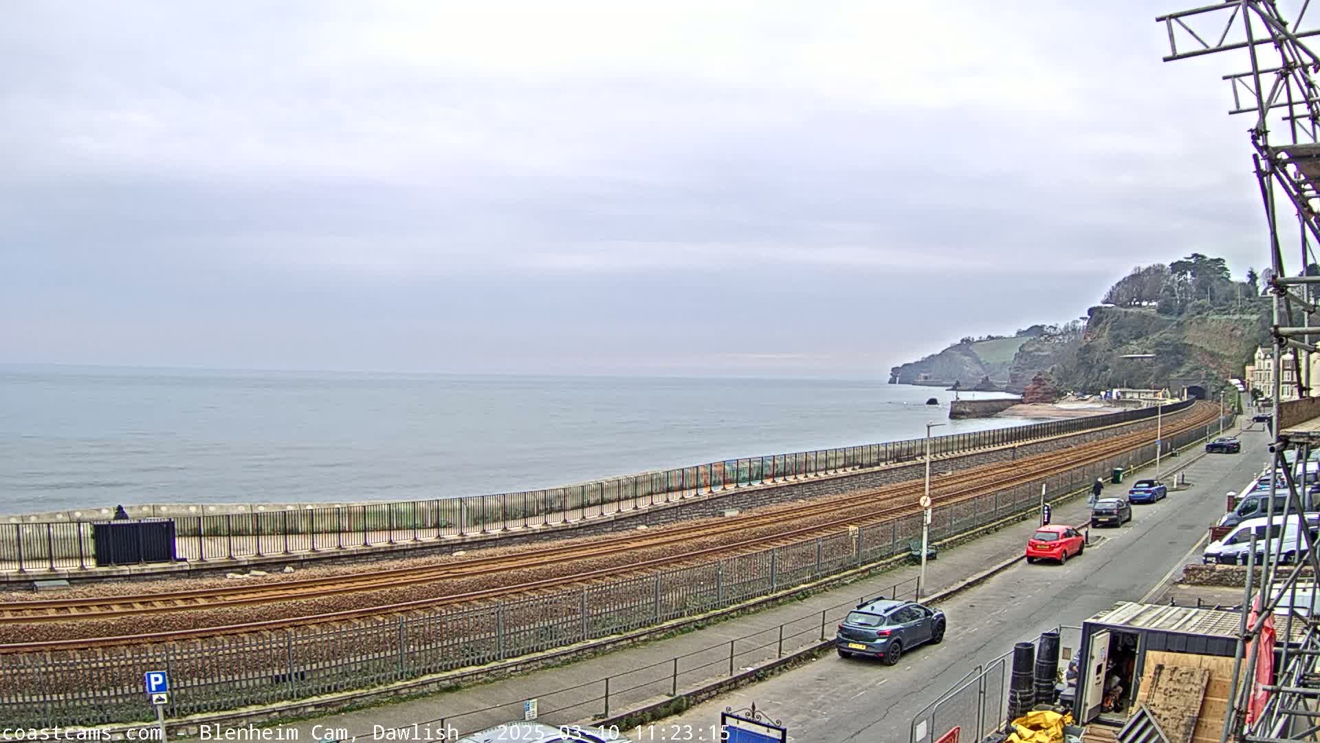A coastal scene under an overcast sky shows train tracks running alongside a seawall separating a beach and the ocean, with cars parked on a street overlooking the scene.
