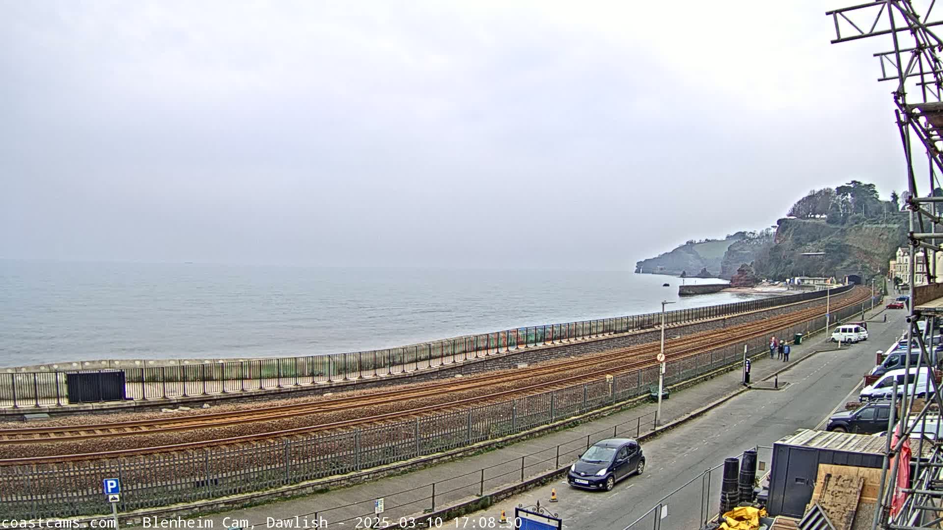 A coastal railway line runs alongside a sea wall, with cars parked on a street overlooking a calm, gray sea and a rocky cliff face under an overcast sky.