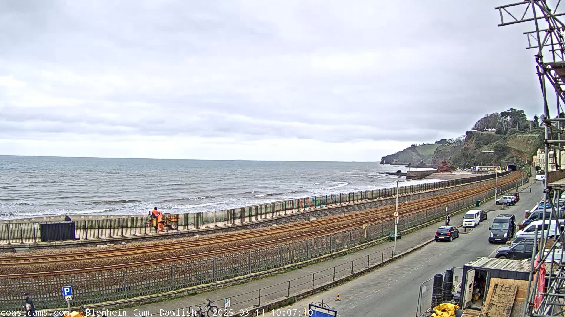 A coastal scene on an overcast day shows railroad tracks running alongside a seawall next to a beach with gentle waves, and a street with parked cars beyond.