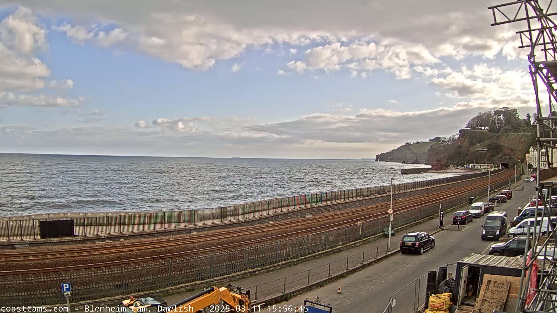 A partly cloudy day shows a coastal scene with a train track running alongside a seawall, several parked cars on a road, and a small, rocky cliff in the distance.